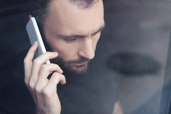 Upset handsome man standing by window with smartphone and looking away — Stock Photo