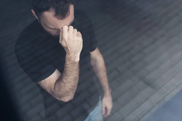 Vue grand angle de l'homme déprimé en t-shirt noir debout près de la fenêtre et tenant la main près du visage — Photo de stock