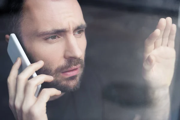 Sad man with smartphone holding hand on window glass and looking away — Stock Photo