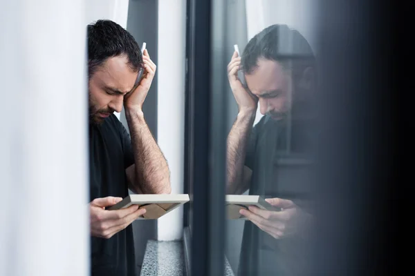 Enfoque selectivo del hombre llorando sosteniendo el marco de la foto mientras está de pie por la ventana - foto de stock