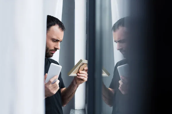 Selective focus of depressed handsome man holding smartphone and photo frame while standing by window — Stock Photo