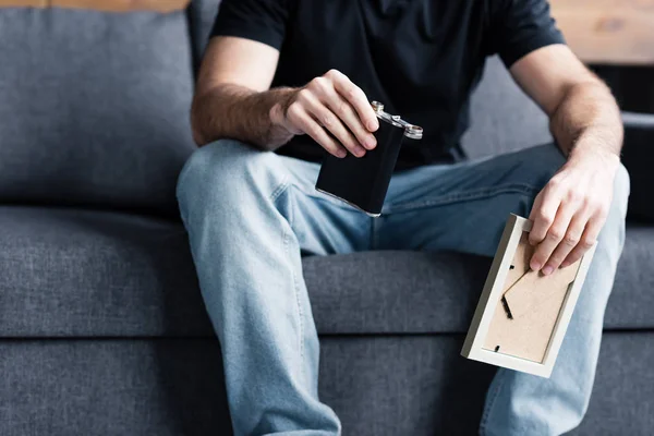 Partial view of man sitting on grey sofa and holding photo frame and flask with alcohol — Stock Photo