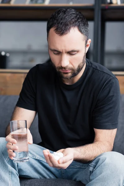 Депресивний бородатий чоловік тримає склянку води і жменьку таблеток — стокове фото