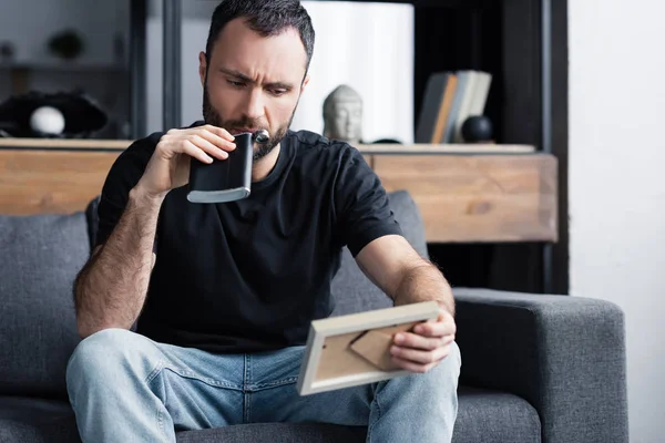 Depressed man drinking from flask and holding photo frame while sitting on sofa at home — Stock Photo