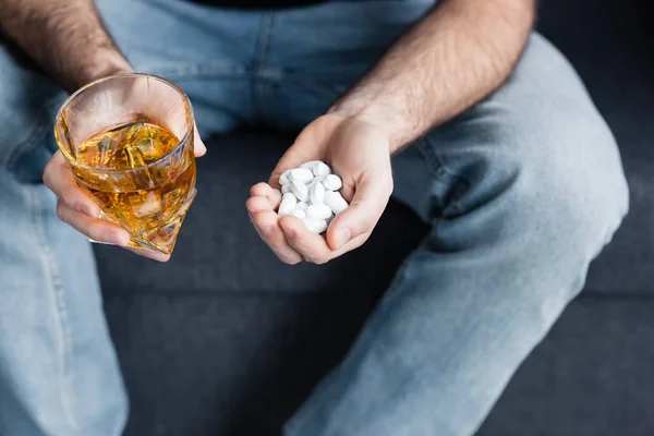 Vista recortada de sentado hombre sosteniendo vaso de whisky y un puñado de pastillas - foto de stock