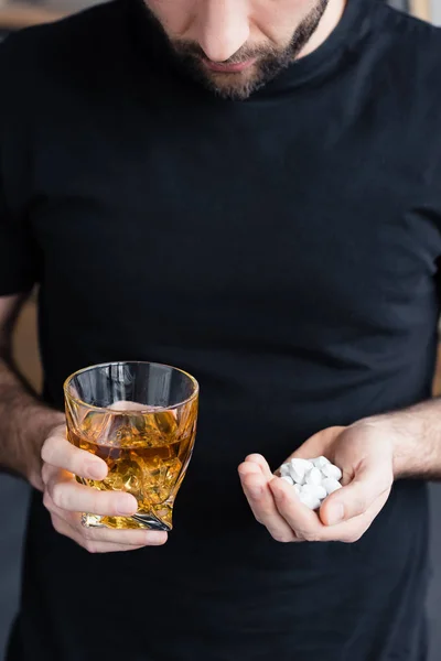 Partial view of standing man in black t-shirt holding glass of whiskey and handful of pills — Stock Photo