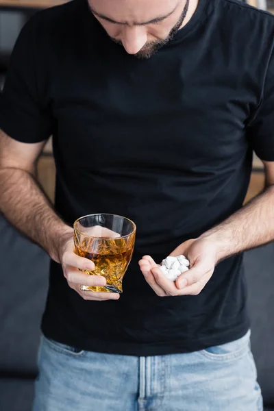 Vista ritagliata di uomo depresso in t-shirt nera con manciata e pillole e bicchiere di whisky — Stock Photo