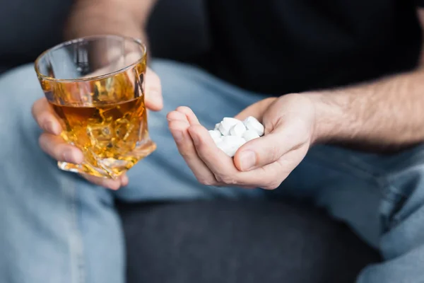 Vue recadrée de l'homme assis avec une poignée de pilules et un verre de whisky — Photo de stock