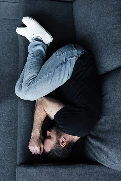 Vista superior del hombre deprimido en camiseta negra y pantalones vaqueros azules acostado en el sofá con los ojos cerrados - foto de stock