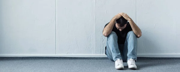 Panoramic shot of depressed man sitting on floor near white wall and holding hands on head — Stock Photo