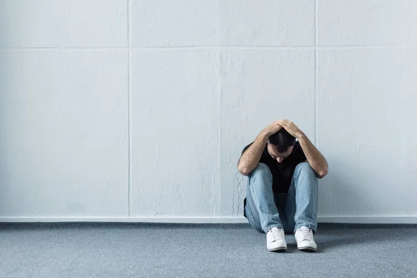 Hombre deprimido sentado en el suelo cerca de la pared blanca y cogido de la mano en la cabeza - foto de stock