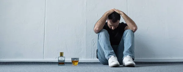 Panoramic shot of suffering man sitting on floor by white wall near bottle and glass of whiskey — Stock Photo