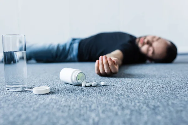 Messa a fuoco selettiva dell'uomo inconscio sdraiato sul pavimento grigio vicino a vetro di acqua e contenitore con pillole — Foto stock