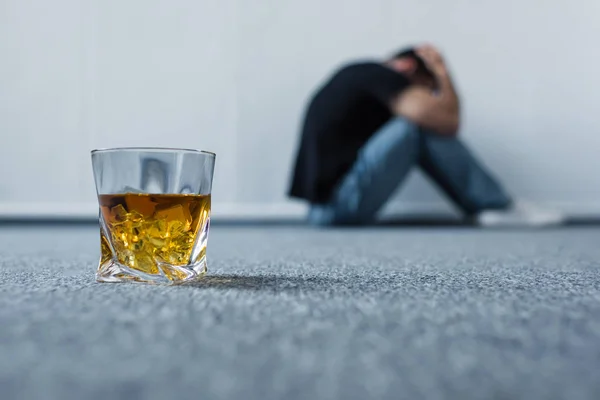 Селективный фокус депрессивного мужчины, страдающего, сидя на сером полу возле стакана виски — стоковое фото