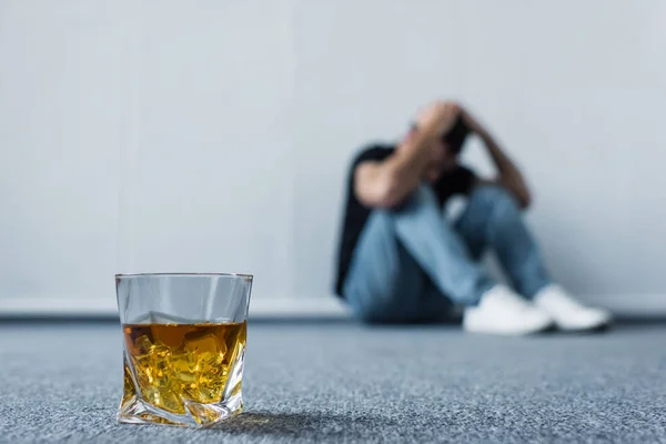 Selektiver Fokus eines depressiven Mannes, der am Boden neben einer weißen Wand in der Nähe eines Whiskeyglases sitzt — Stockfoto