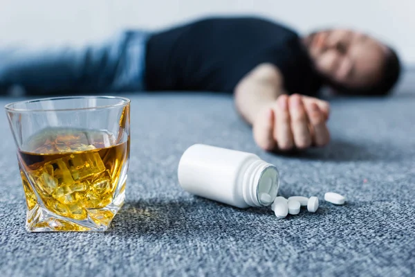 Selective focus of unconscious man lying on floor near glass of whiskey and container with pills — Stock Photo