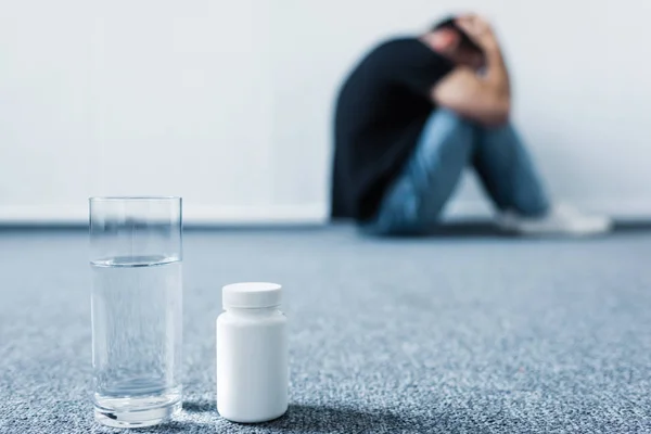 Foco seletivo do homem deprimido sofrendo enquanto sentado no chão por parede perto do recipiente com pílulas e vidro de água — Fotografia de Stock