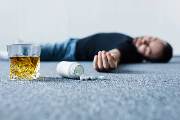 Foco seletivo do homem inconsciente deitado no chão cinza perto de vidro de uísque e recipiente com pílulas — Fotografia de Stock