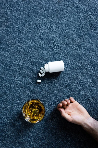 Вид сверху на мужскую руку возле стакана виски и контейнера с таблетками на сером полу — стоковое фото