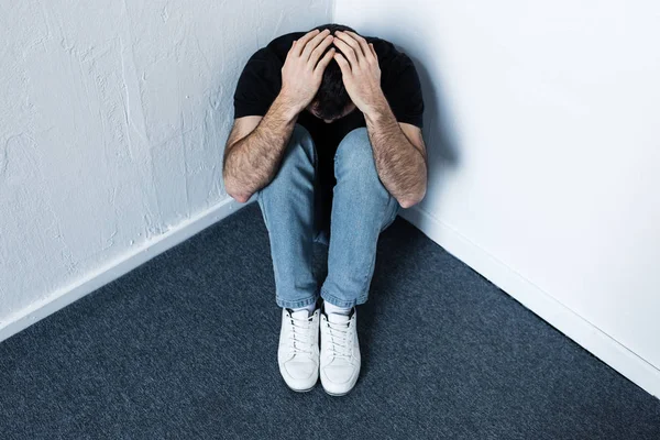 Depressiver Mann sitzt auf grauem Boden in Ecke und hält Hände auf dem Kopf — Stockfoto