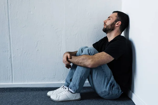 Hombre deprimido guapo sentado en el suelo en esquina por la pared blanca y mirando hacia otro lado - foto de stock