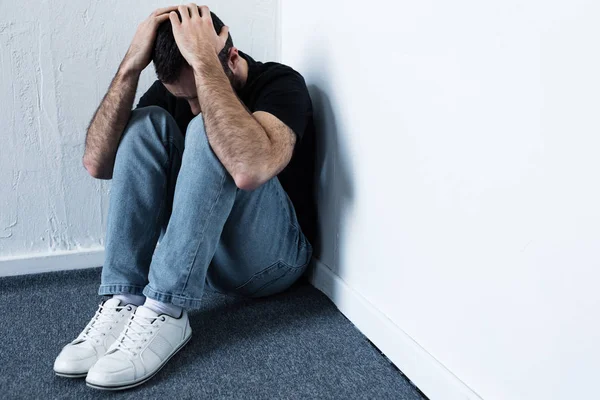 Depressed man in blue jeans and white sneakers sitting in corner and holding hands on head — Stock Photo