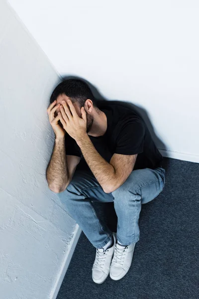 Vista de ángulo alto del hombre deprimido adulto sentado en el suelo en la esquina y cubriendo la cara con las manos - foto de stock