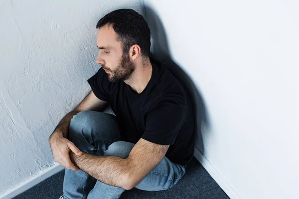 Handsome bearded depressed man sitting on floor in corner and looking away — Stock Photo