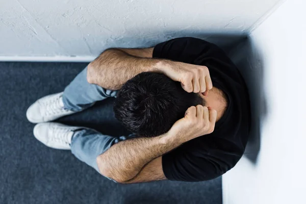 Vista superior del hombre adulto deprimido sentado en el suelo en la esquina y cogido de la mano en la cabeza - foto de stock