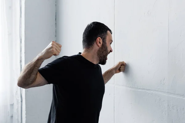 Hombre barbudo enojado en camiseta negra gritando y pateando la pared blanca — Stock Photo