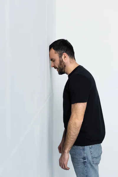 Homme barbu frustré en t-shirt noir debout près du mur blanc avec les yeux fermés — Photo de stock