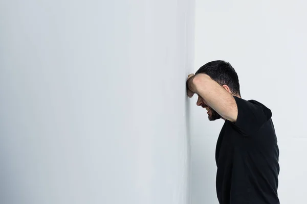 Hombre deprimido en camiseta negra sufriendo mientras está de pie junto a la pared blanca - foto de stock