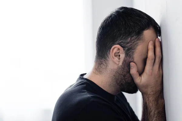 Hombre adulto triste de pie junto a la pared blanca y cubriendo la cara con las manos - foto de stock