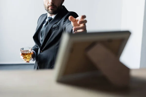 Selective focus of man in black suit holding glass of whiskey and outstretching hand to desk with photo frame — Stock Photo