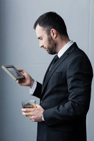 Hombre barbudo deprimido en traje mirando la foto en el marco mientras sostiene el vaso de whisky - foto de stock