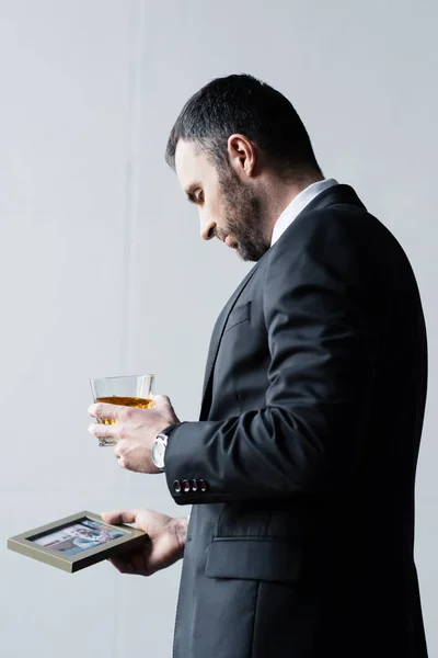 Upset bearded man in suit holding photo in frame and glass of whiskey — Stock Photo