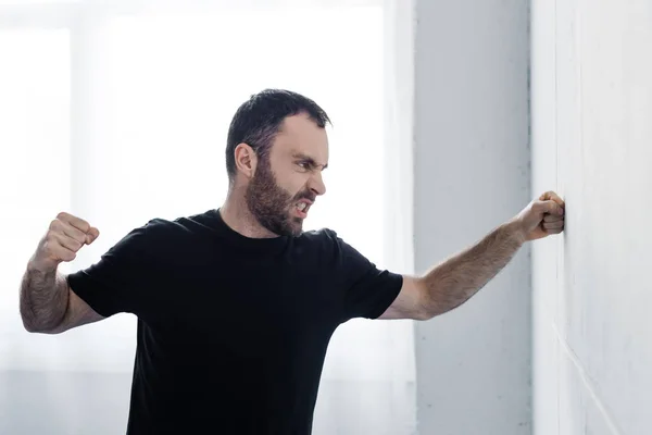 Arrabbiato uomo barbuto in nero t-shirt calci muro bianco con la mano — Foto stock