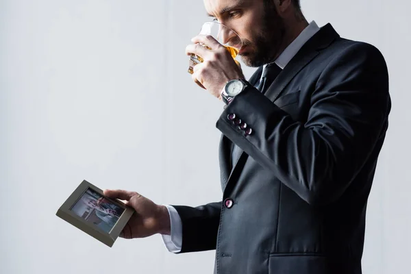 Hombre molesto en traje bebiendo whisky y mirando la foto en el marco - foto de stock
