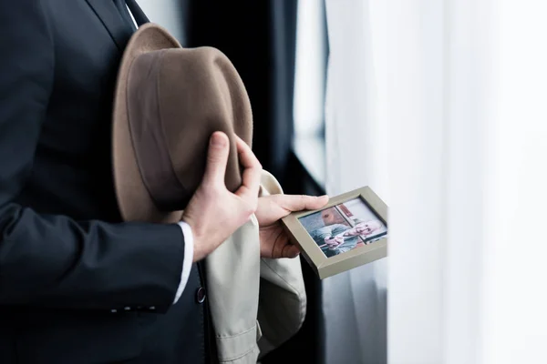 Vista ritagliata di uomo che tiene la foto in cornice mentre in piedi dalla finestra con il cappello in mano — Foto stock