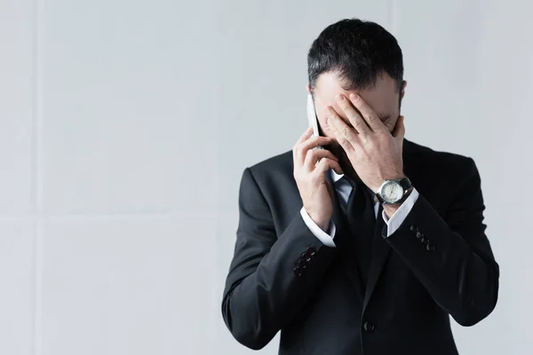 Hombre decepcionado en traje negro usando teléfono inteligente y sosteniendo la mano en la cara - foto de stock