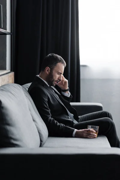 Aufgebrachter Geschäftsmann im Anzug sitzt mit Glas Whiskey auf Sofa und benutzt Smartphone — Stockfoto