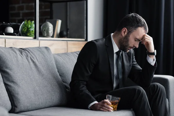 Hombre frustrado en traje sentado en el sofá con los ojos cerrados y sosteniendo vaso de whisky - foto de stock