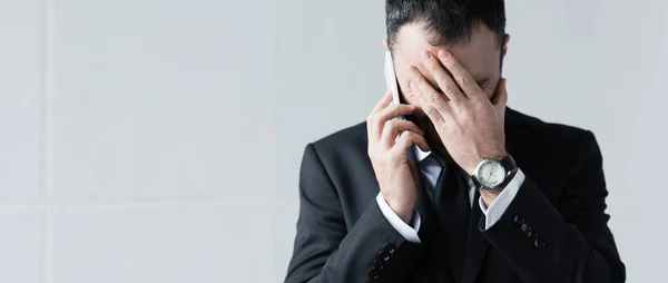 Панорамный снимок расстроенного бизнесмена, держащегося за лицо во время использования смартфона — стоковое фото