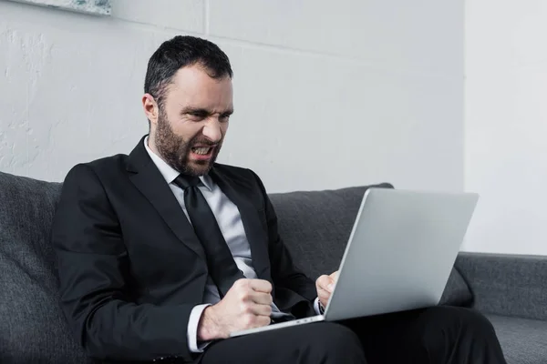 Сердитий бізнесмен з руками в кулаках, використовуючи ноутбук, сидячи на дивані в офісі — стокове фото