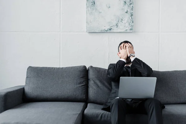 Расстроенный бизнесмен держит руки на лице, сидя на сером диване с ноутбуком — стоковое фото