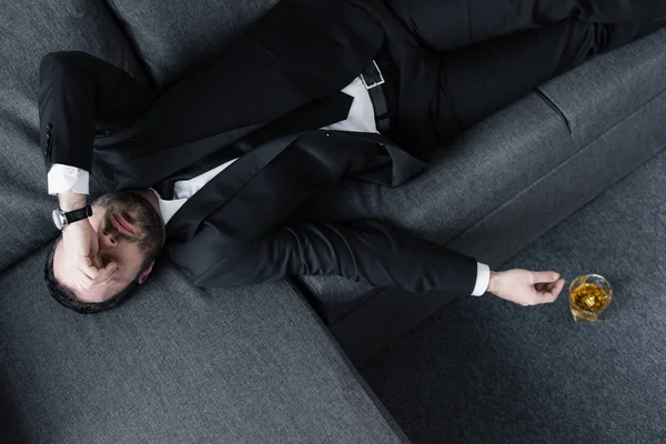 Top view of depressed businessman lying on sofa near glass of whiskey on floor — Stock Photo