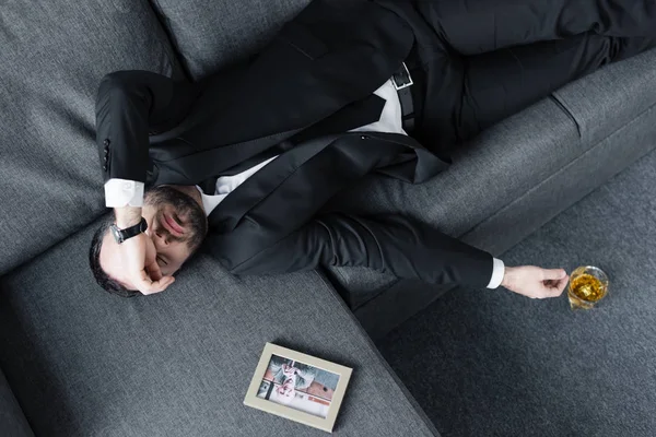 Draufsicht eines frustrierten Geschäftsmannes, der auf einem Sofa neben einem Foto im Rahmen liegt und ein Glas Whiskey auf dem Boden — Stockfoto