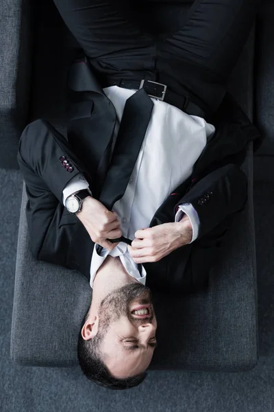 Vista superior de hombre de negocios deprimido en traje negro que sufre mientras está acostado en el sofá - foto de stock