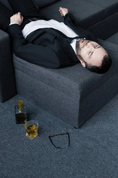 High angle view of upset businessman lying on sofa near glasses, bottle and glass of whiskey on floor — Stock Photo