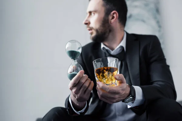 Избирательный фокус разочарованного бизнесмена, держащего стакан виски и песочные часы — стоковое фото